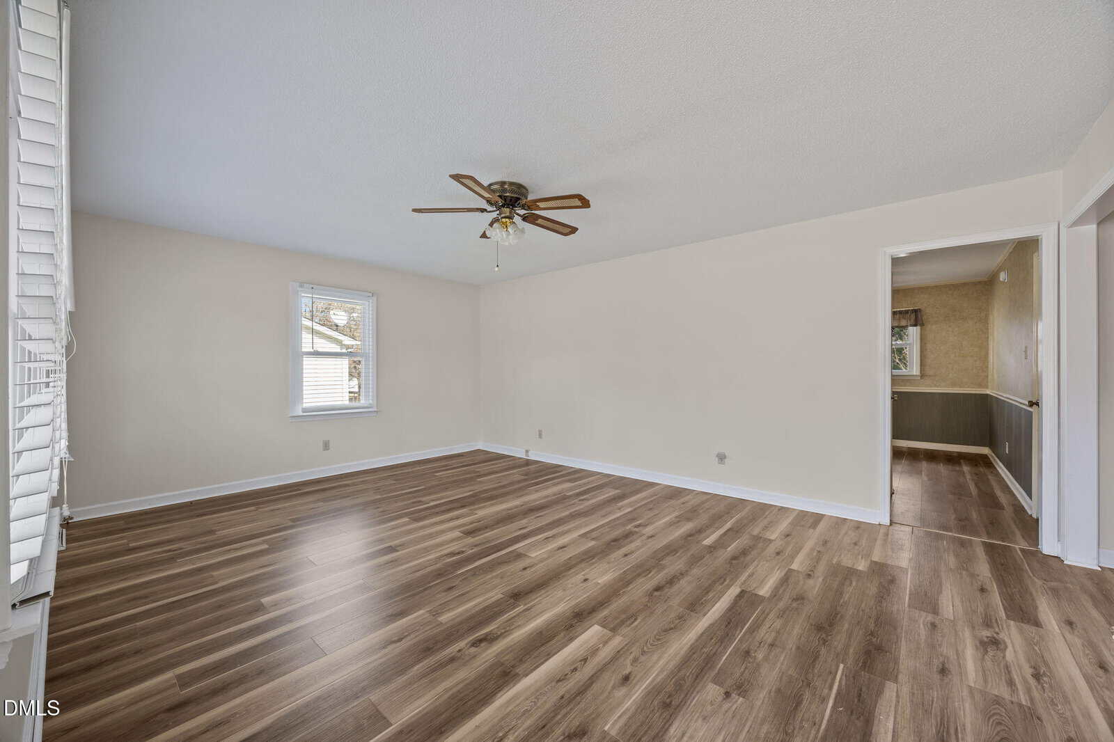 Empty room with beige walls, wood-look laminate flooring, a ceiling fan with lights, two windows with white blinds, and an open doorway leading to another space. Natural light enters from the windows.