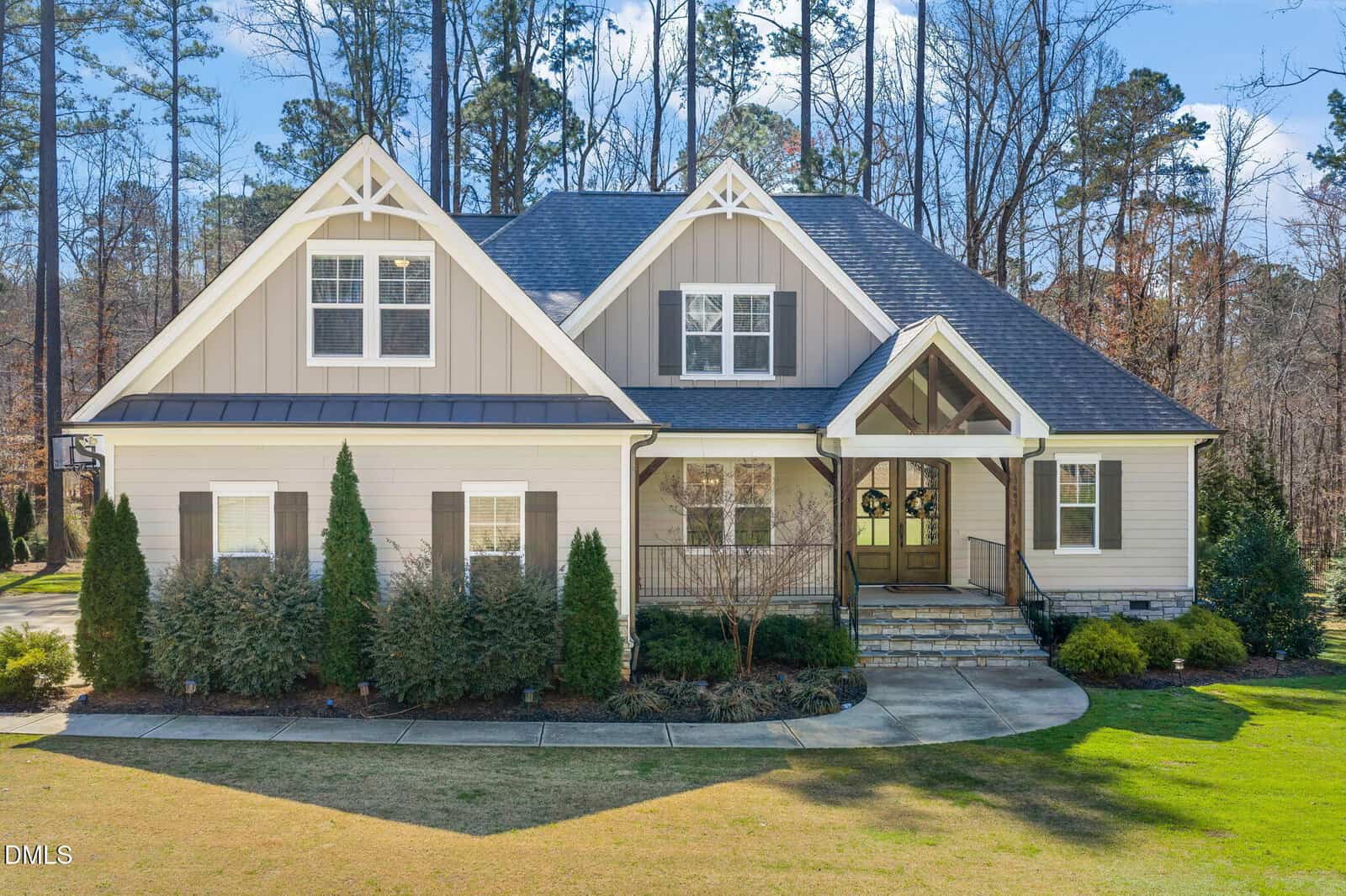 A two-story house with beige siding, white trim, peaked roofs, and large front windows is nestled on 3603 Carole Court in Wake Forest. Shrubs line the front of the home, and a curved walkway leads to double front doors, set against a beautiful backdrop of tall trees.