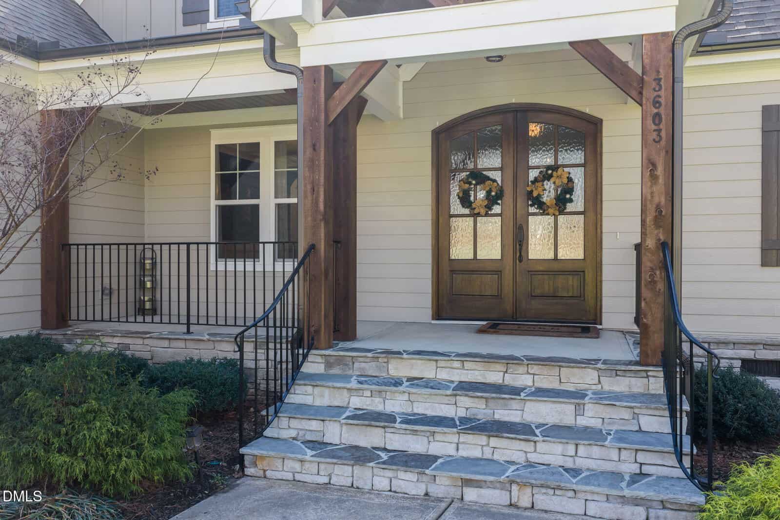 Front entrance of a house with stone steps, a double wooden door decorated with wreaths, beige siding, black metal railings, and some shrubs and plants near the walkway. House number 3603 is visible on the right post.