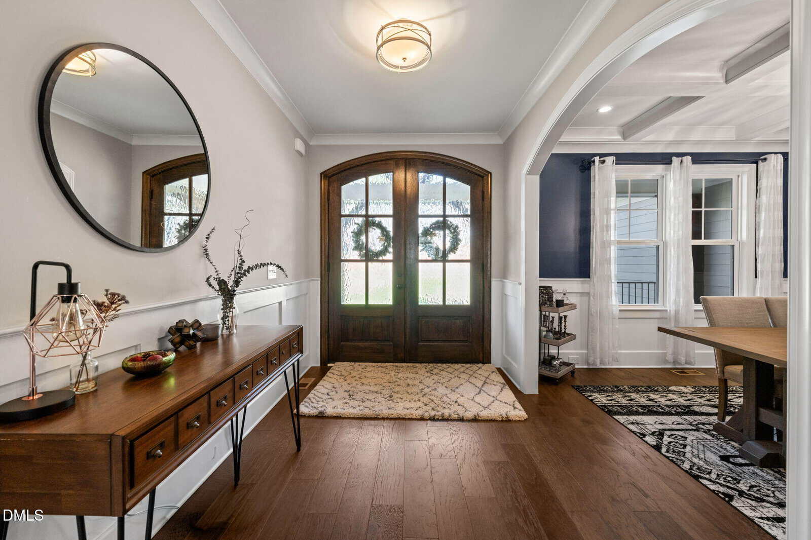 A foyer with wooden double doors, a patterned rug, a wooden console table with decor items, a round mirror, and an archway leading to a dining room with a large window and white curtains.