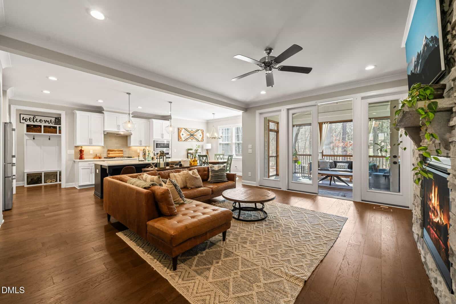 Open-concept living room and kitchen with brown sectional sofa, fireplace, large rug, ceiling fan, and glass doors leading to a screened porch. Kitchen features white cabinets, island, and stainless steel appliances.