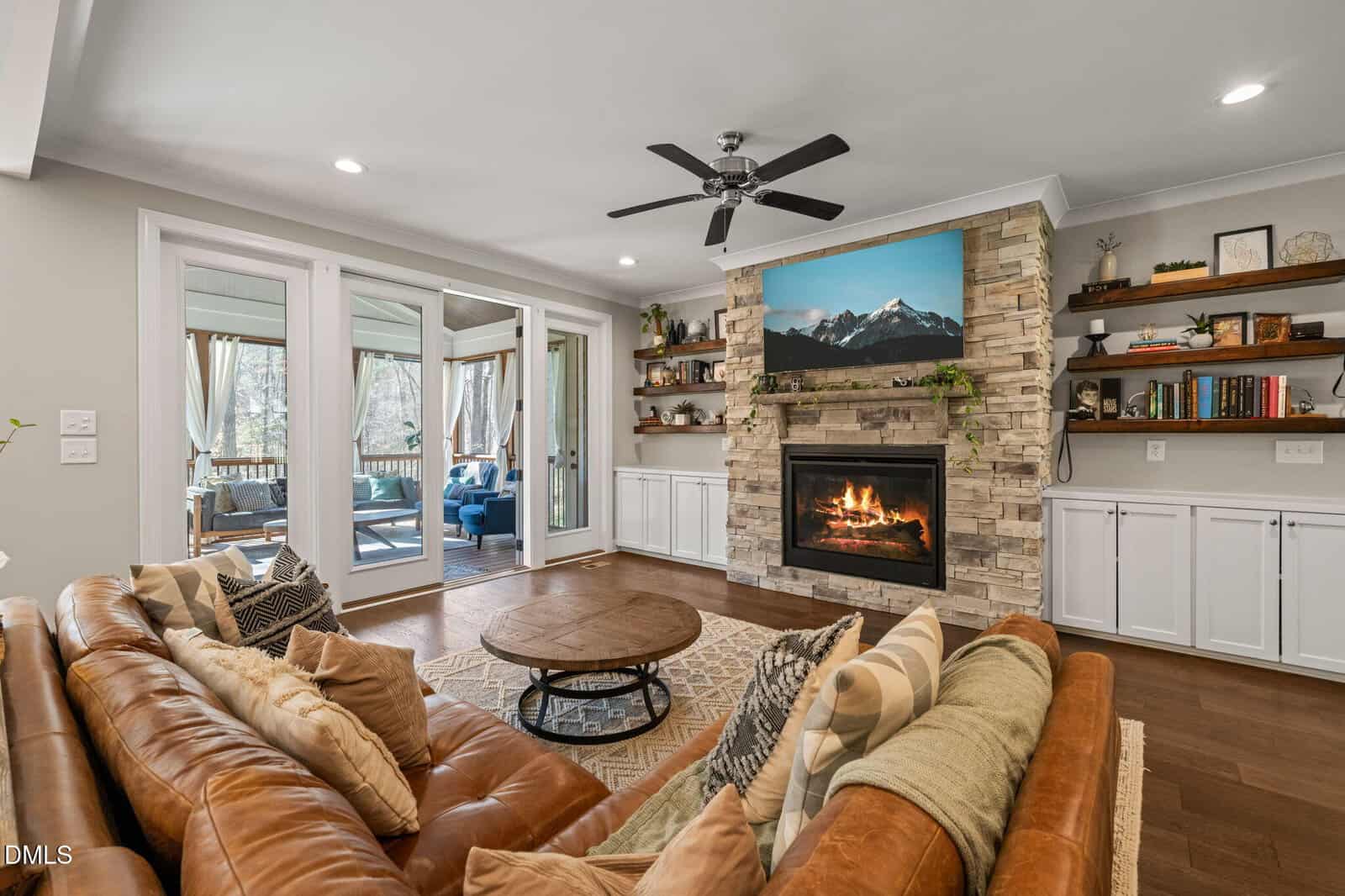 A living room with a leather sofa, round wooden coffee table, stone fireplace with a mountain photo above, built-in shelves, cabinets, and sliding glass doors leading to a covered patio with outdoor seating.