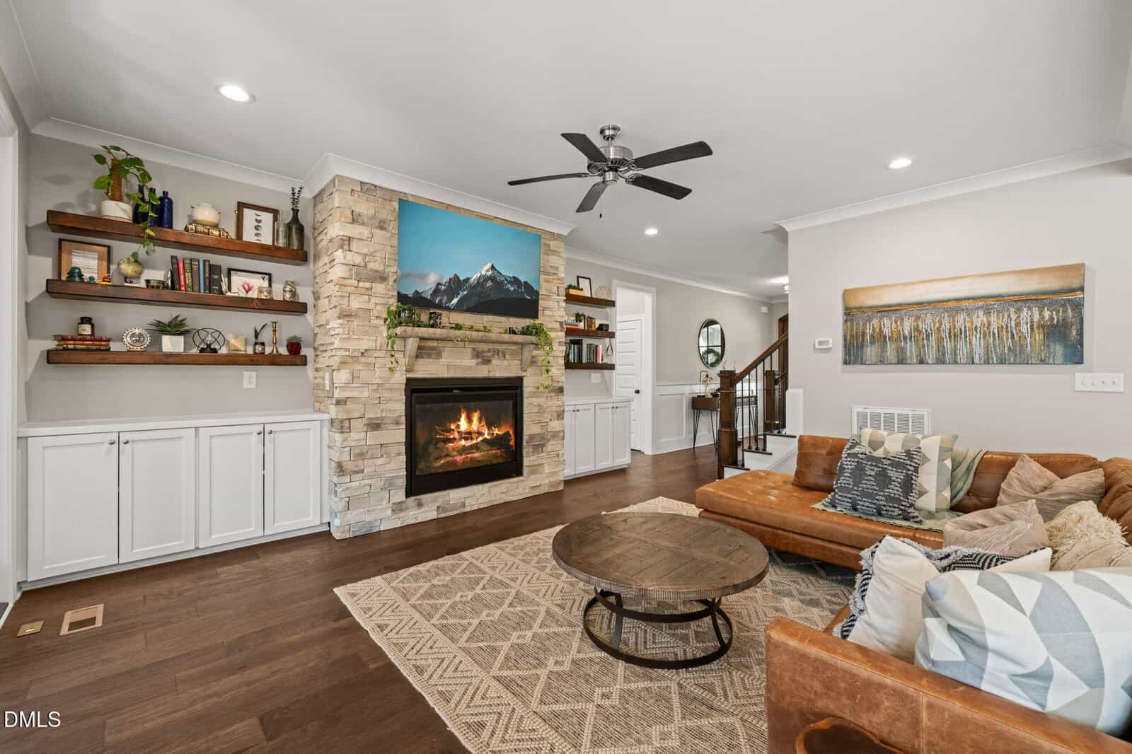 A cozy living room with a stone fireplace, a brown sectional sofa, built-in shelves, a round coffee table, and a patterned rug. A TV above the fireplace displays a mountain scene.
