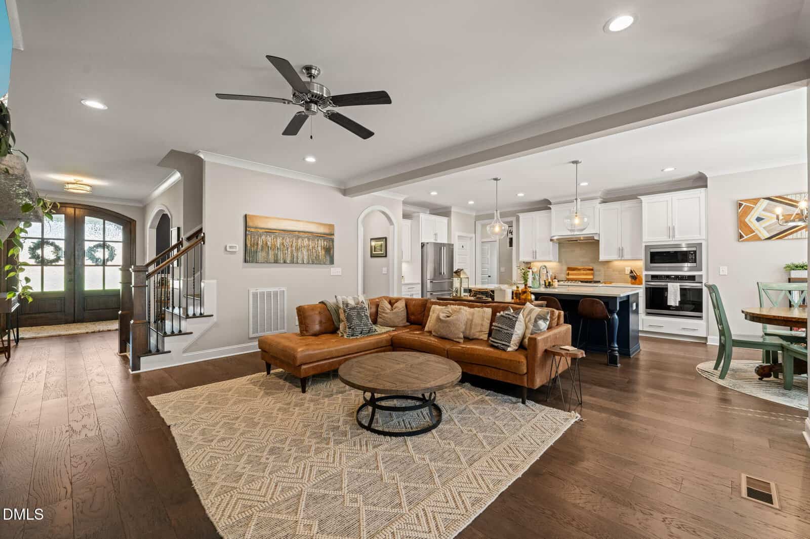 Open-concept living space with a brown sectional sofa, round coffee table, patterned rug, modern kitchen with island, and dining area; wooden floors, ceiling fan, and recessed lights.