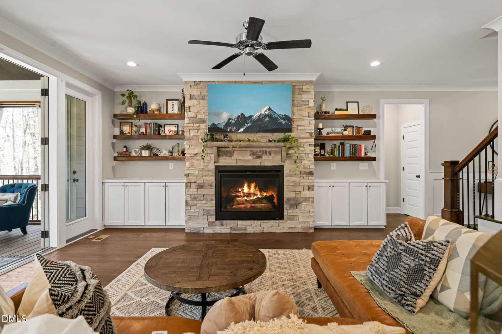 A cozy living room with a stone fireplace, fire burning, a TV above showing mountains, built-in shelves on both sides, a ceiling fan, and a brown sofa with patterned pillows in the foreground.