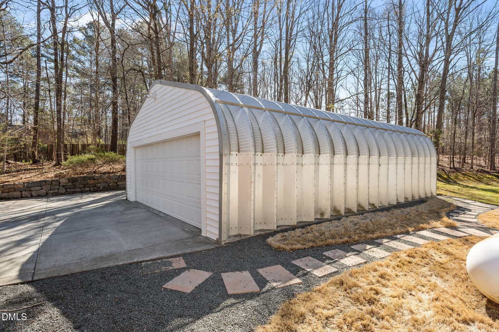 A white metal Quonset hut-style garage with a double door sits on a concrete driveway, surrounded by dry grass and leafless trees under a clear blue sky.