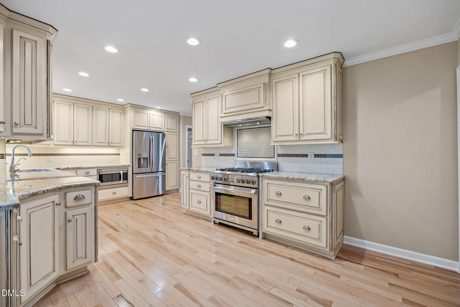 Spacious kitchen with cream cabinets, stainless steel appliances, gas range, granite countertops, light wood floors, and recessed lighting. Neutral wall color and decorative tile backsplash above the stove.