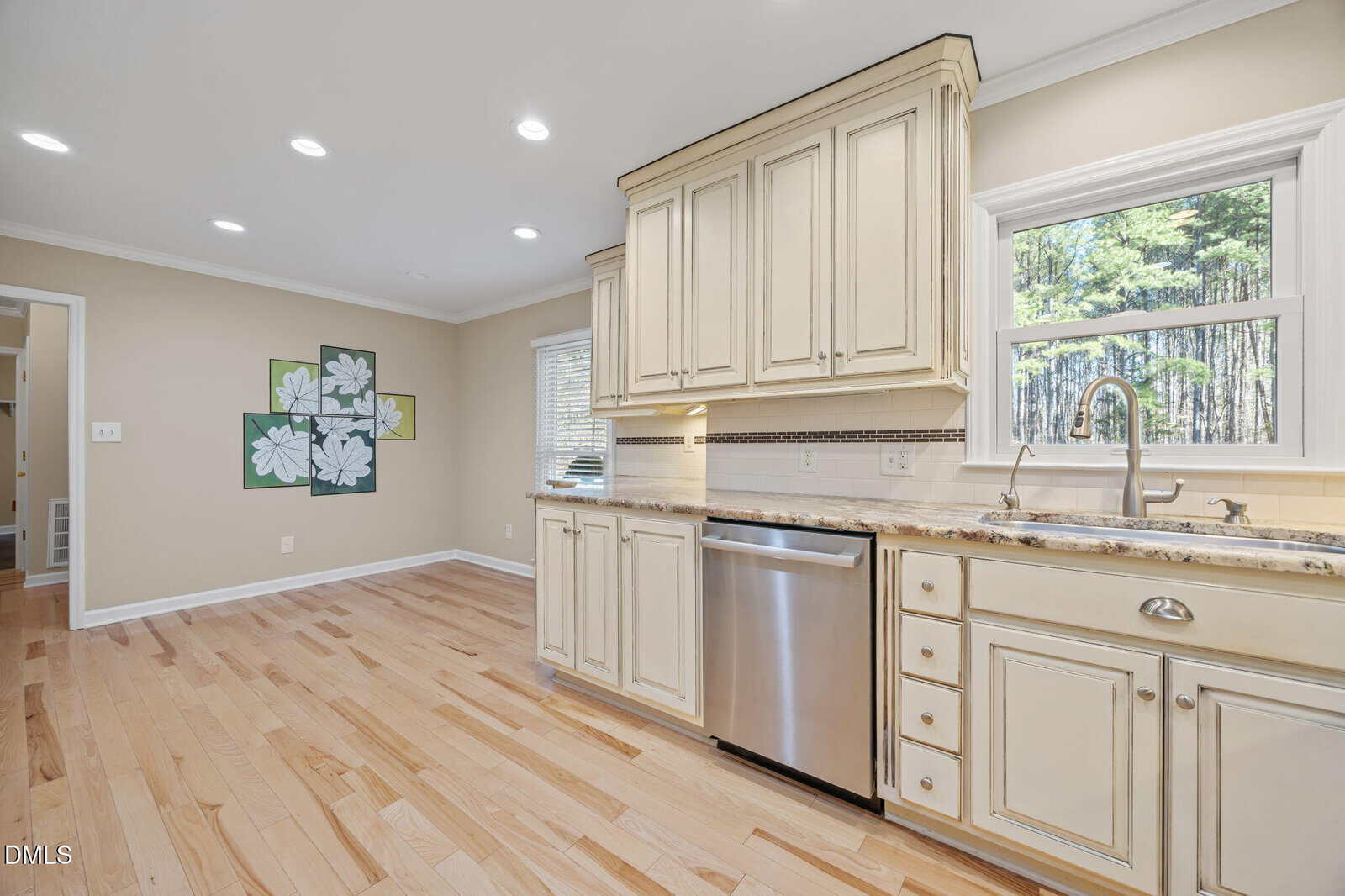 Bright kitchen with cream cabinets, stainless steel dishwasher, marble countertops, and light wood floors. Window above sink shows trees. Adjacent wall features a floral art piece and leads to another room.