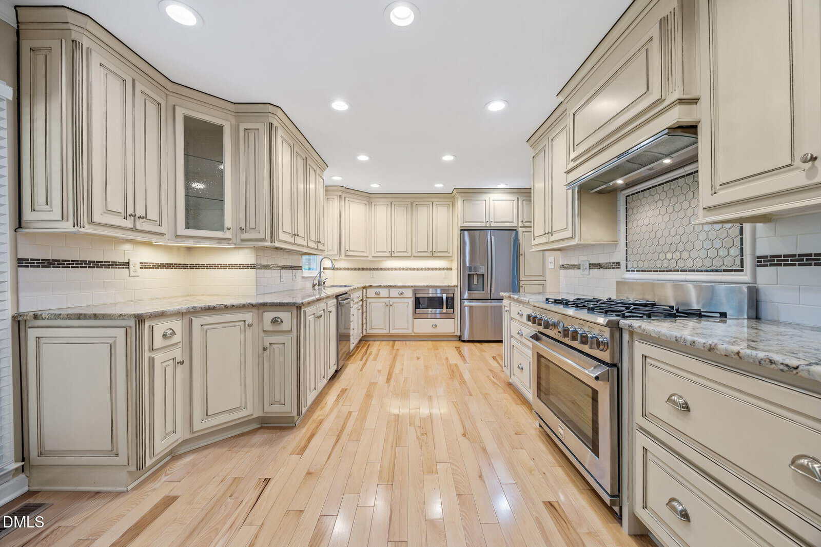 Spacious kitchen with light wood floors, cream-colored cabinets, granite countertops, stainless steel appliances, a large gas stove, and a built-in refrigerator. Recessed lighting brightens the room.