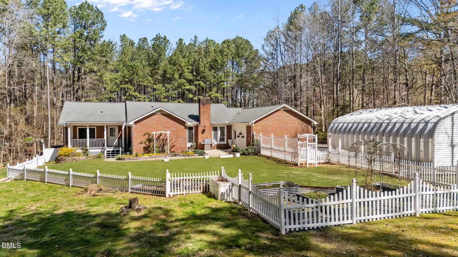 A red brick house with a gray roof sits in a large yard, surrounded by white picket fencing and trees. A greenhouse structure is on the right, with garden beds and a manicured lawn in front of the house.
