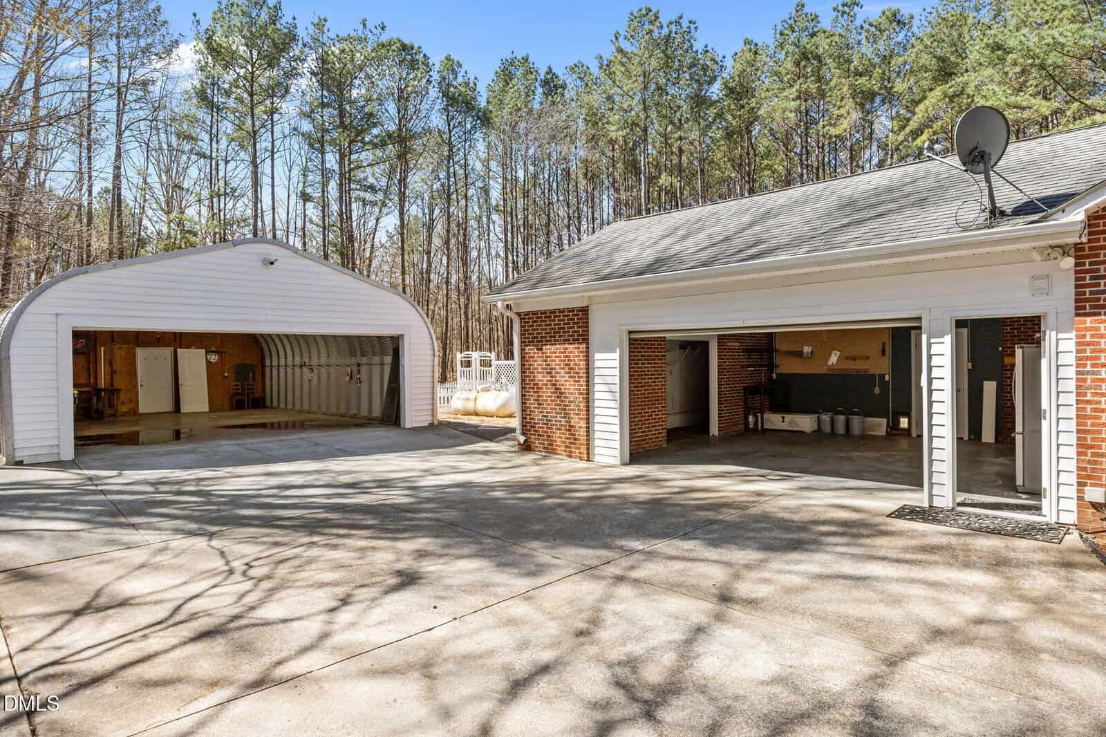 A driveway leads to a brick house with two open garage doors and a detached white metal storage building, surrounded by tall trees under a clear blue sky.