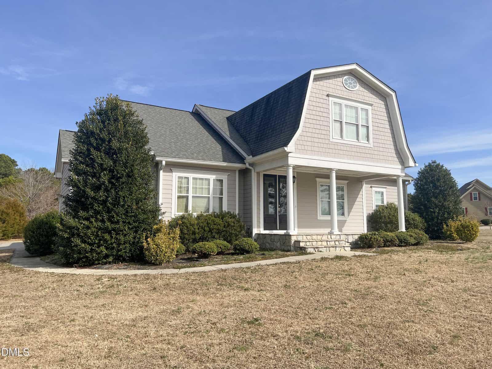 A beige two-story house with a gambrel roof, white trim, front porch, and shrubbery in the yard, set against a blue sky with some neighboring houses visible in the background.