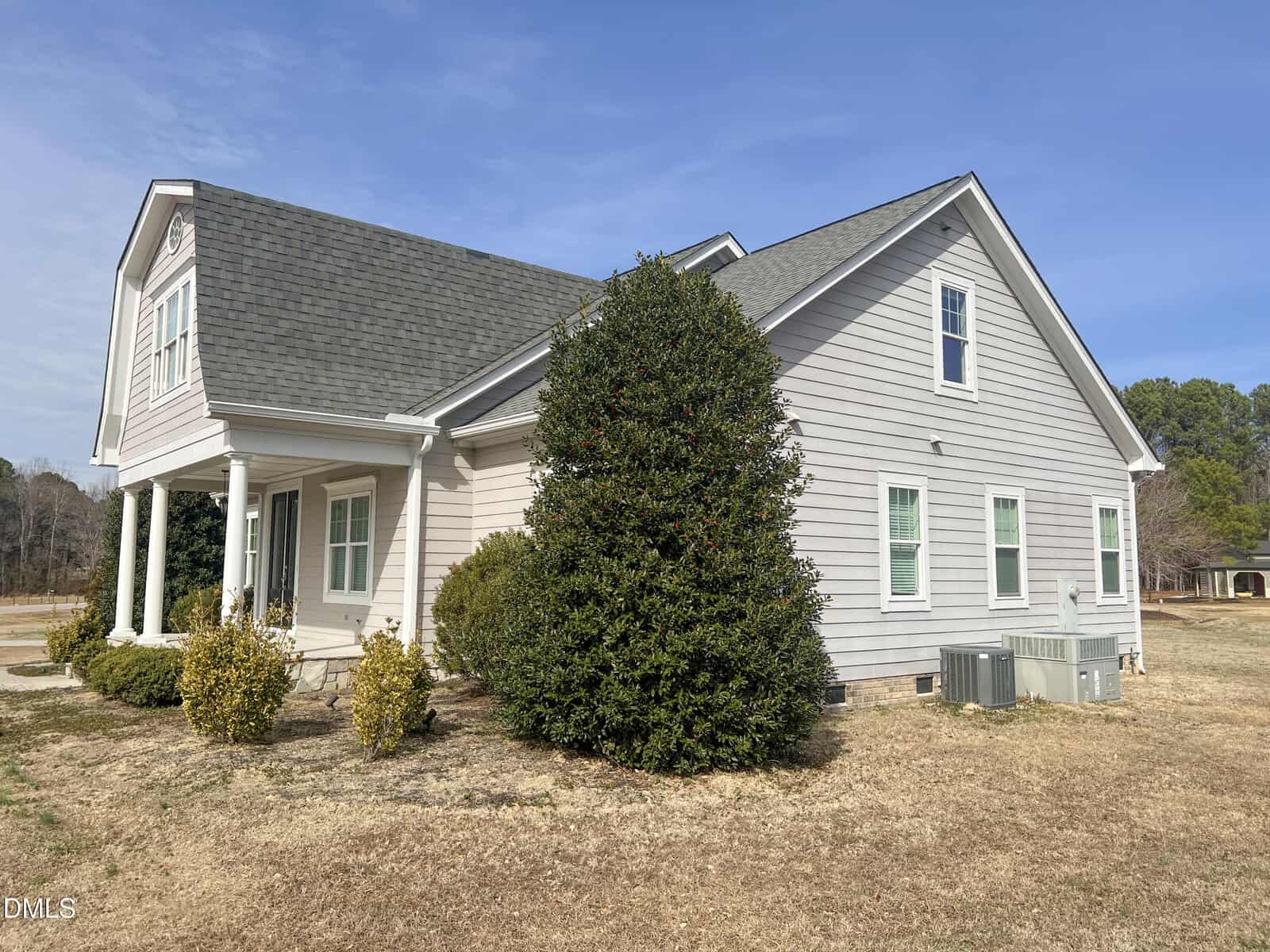 A light gray, single-story house with white trim and a gabled roof. Several bushes and a large shrub are in front. Two air conditioning units are on the right side. The lawn is dry and patchy, and the sky is clear.