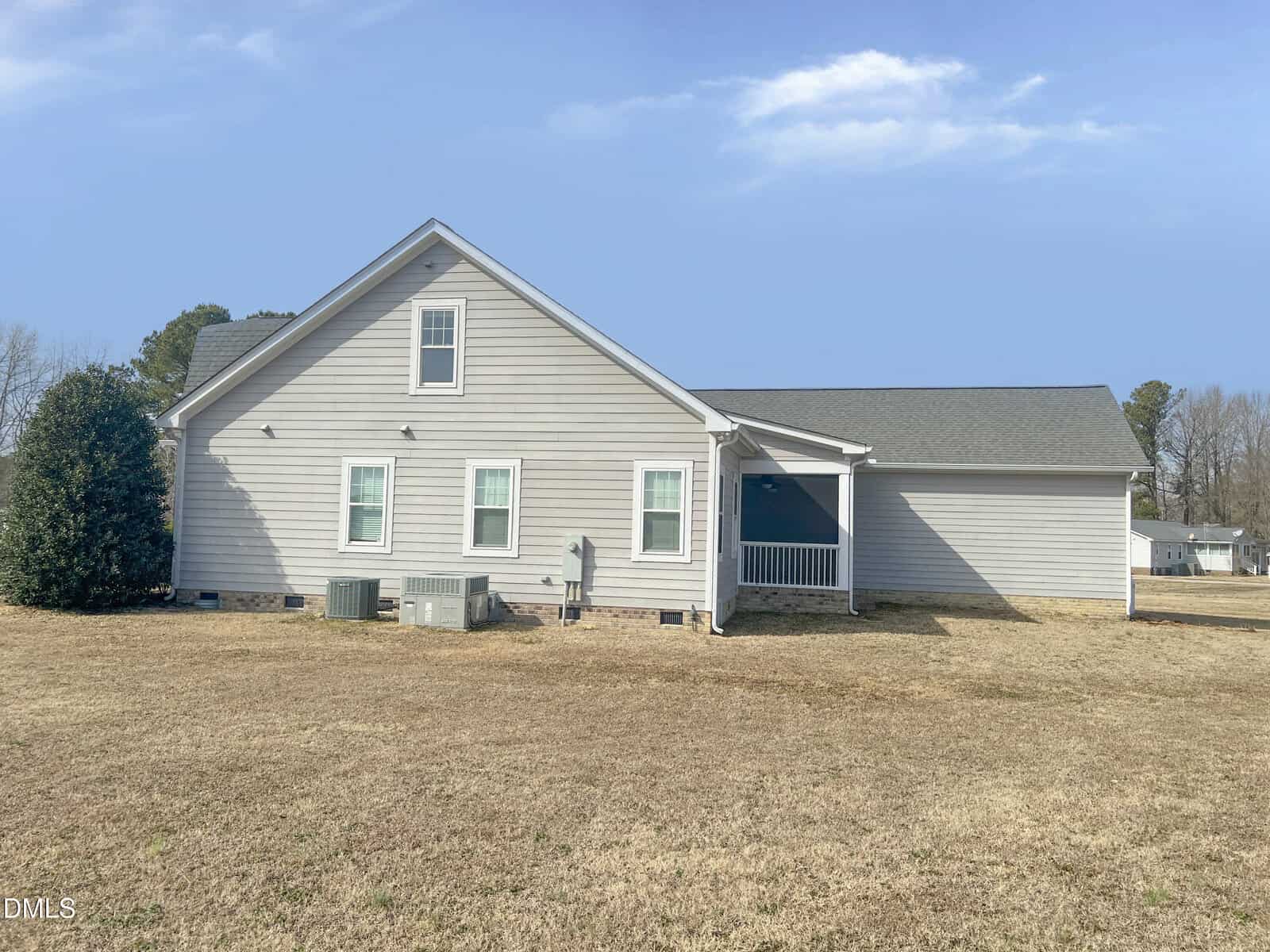 Single-story light gray house with white trim, two visible windows, side porch, and HVAC units on a dry, patchy lawn. The sky is clear with a few clouds and trees are in the background.