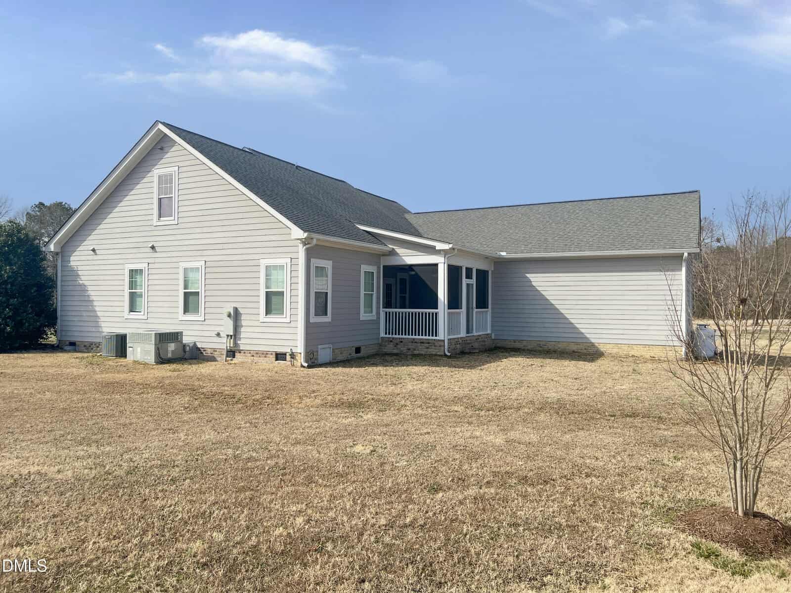 A single-story gray house with white trim, a gable roof, several windows, a small screened porch, and an attached garage sits on a dry, brown grassy yard under a partly cloudy sky.