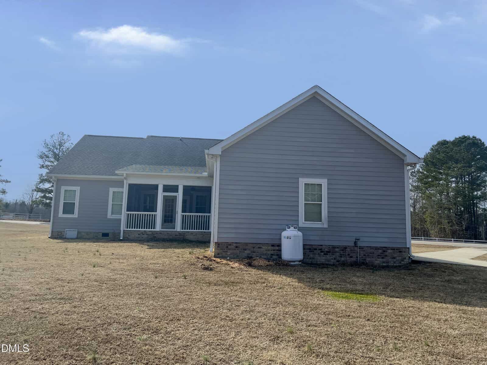 A single-story house with gray siding, white trim, a covered back porch, and a propane tank sits on a large lot with sparse grass under a clear blue sky. Trees line the background.