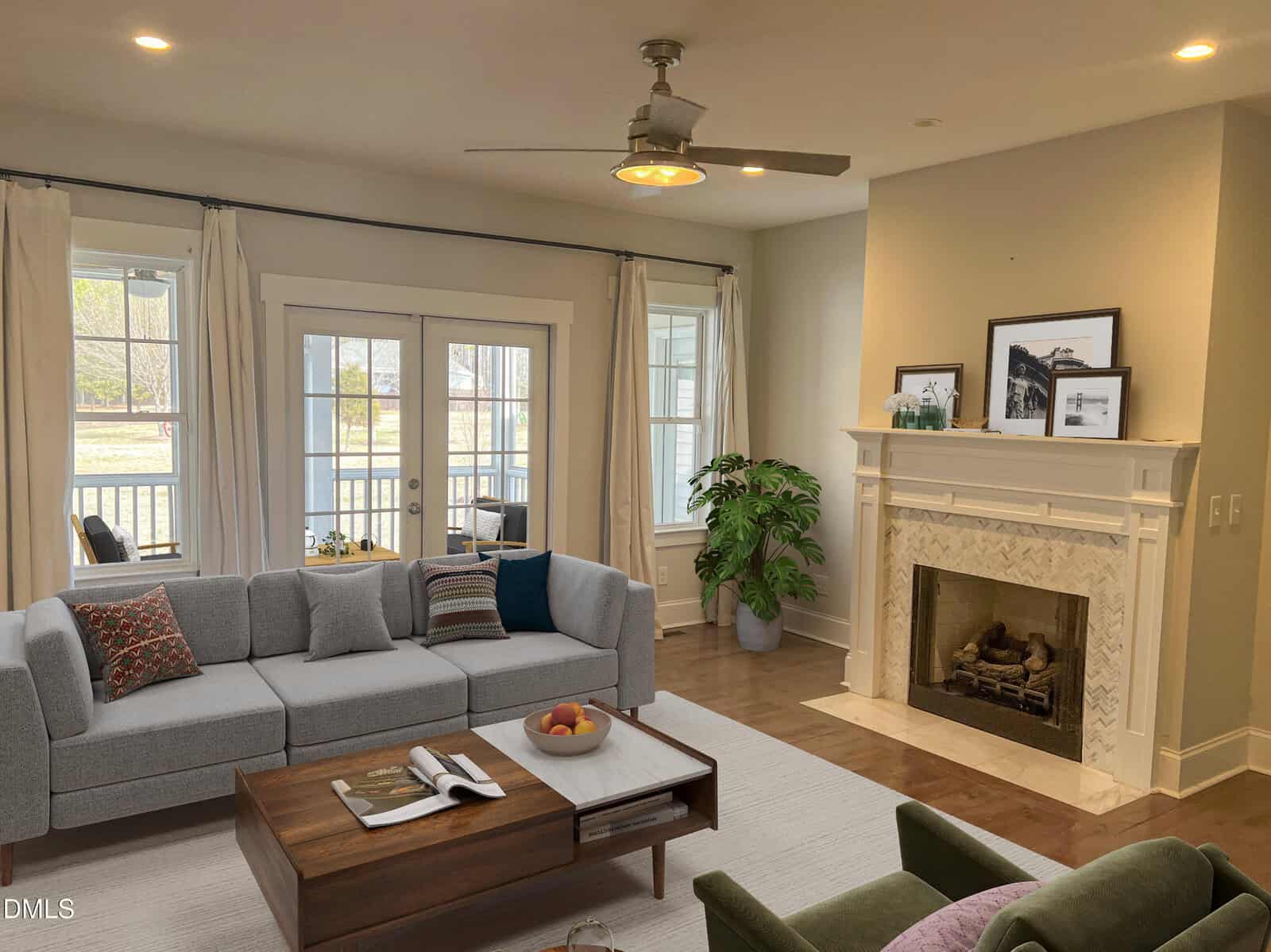 A bright living room with a gray sectional sofa, wooden coffee table, fireplace with a white mantel, framed art, potted plant, and glass doors leading to a sunroom. Light walls and wood floors create a cozy atmosphere.