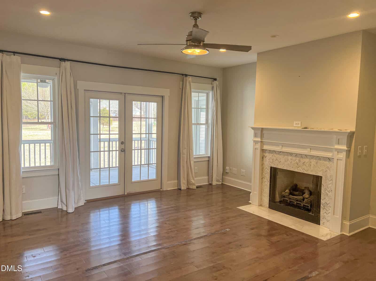 A living room with hardwood floors, a ceiling fan with light, white curtains on windows and French doors, and a white fireplace with a marble surround. The room has neutral-colored walls and natural light.