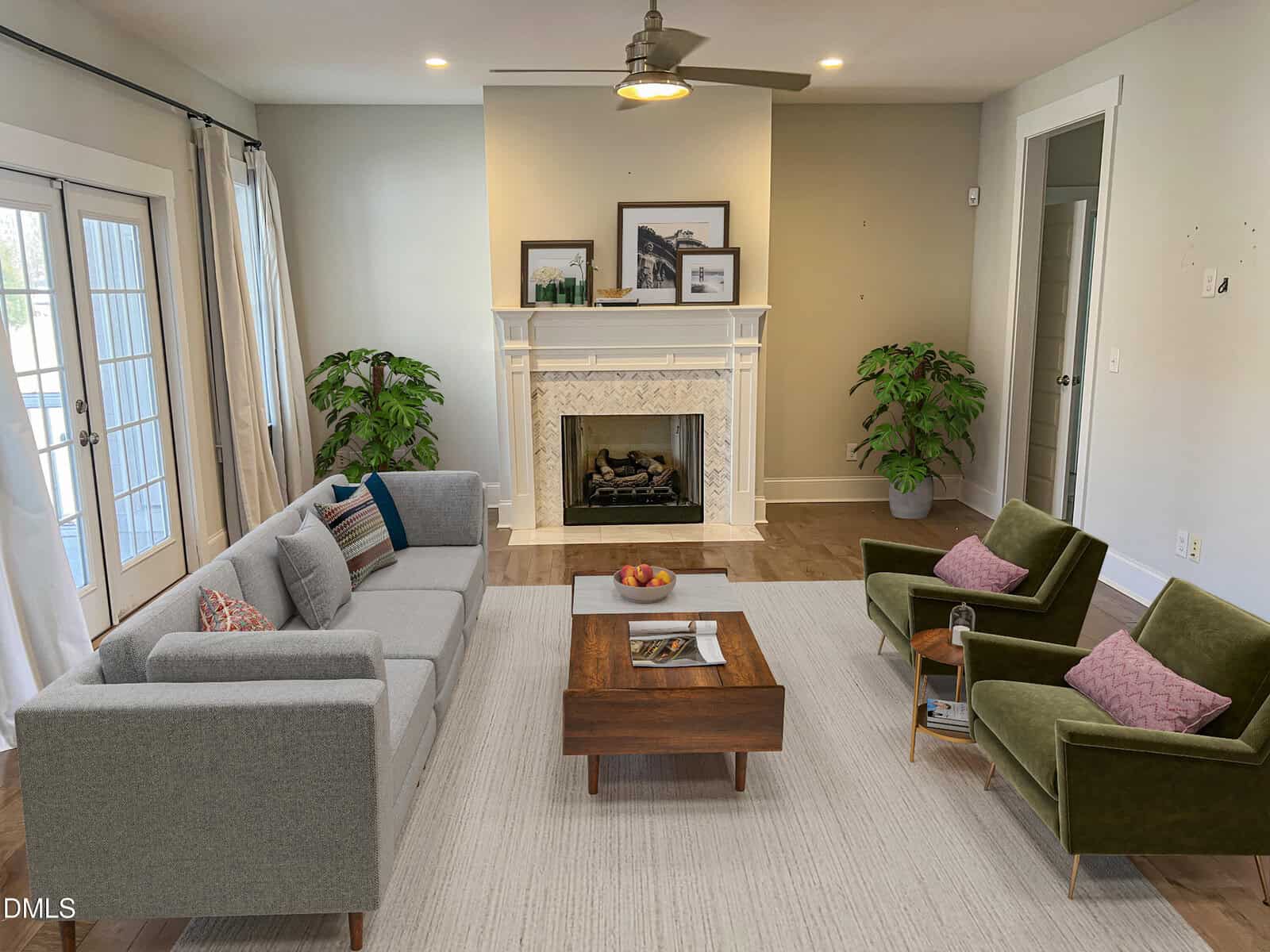 A modern living room with a gray sofa, two green armchairs, a wooden coffee table, a white fireplace, framed art above the mantel, two potted plants, large windows with sheer curtains, and a light rug on a wood floor.