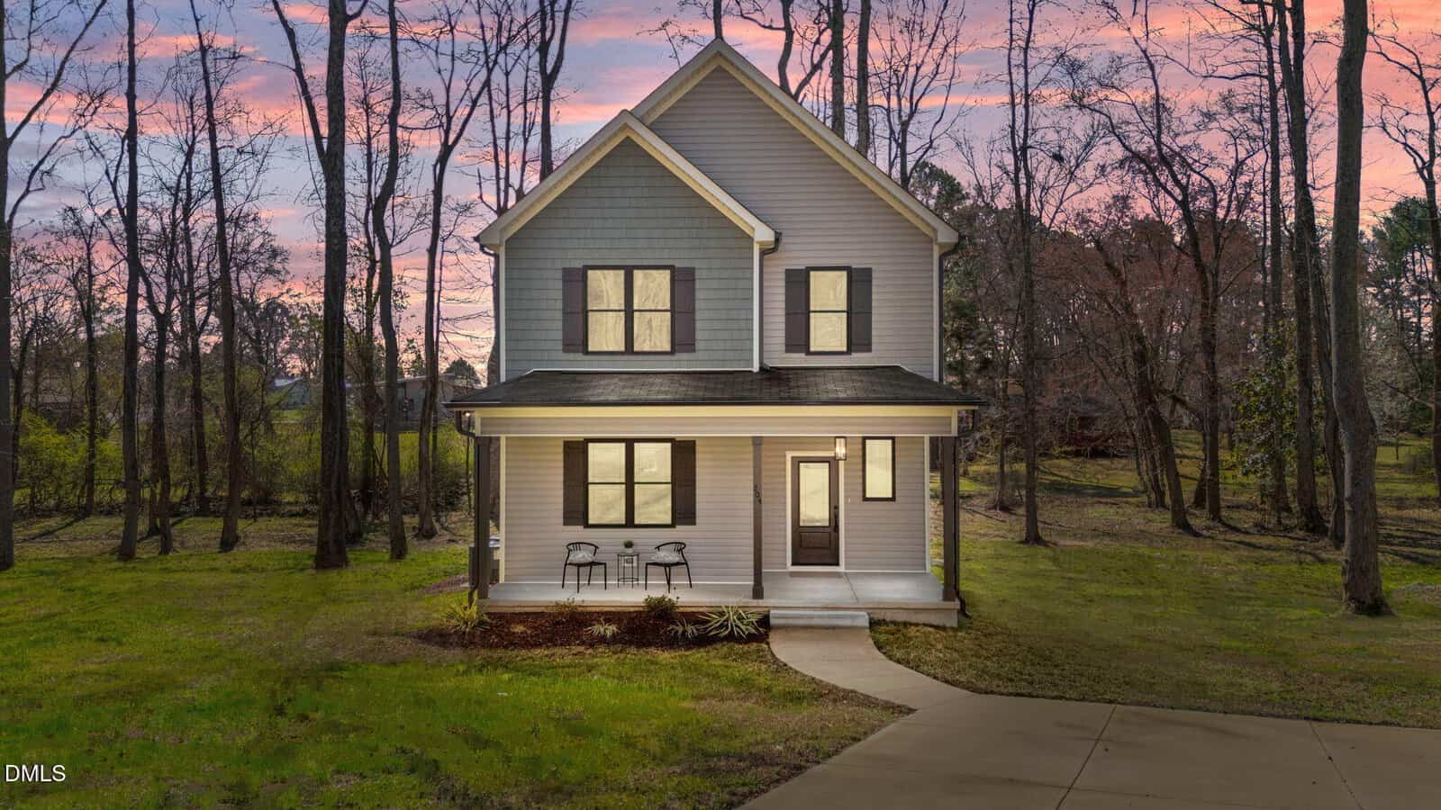 A two-story gray house with white trim and a covered front porch, featuring two chairs and a table. The house sits on a grassy lot surrounded by trees under a sunset sky. A concrete driveway is in the foreground.