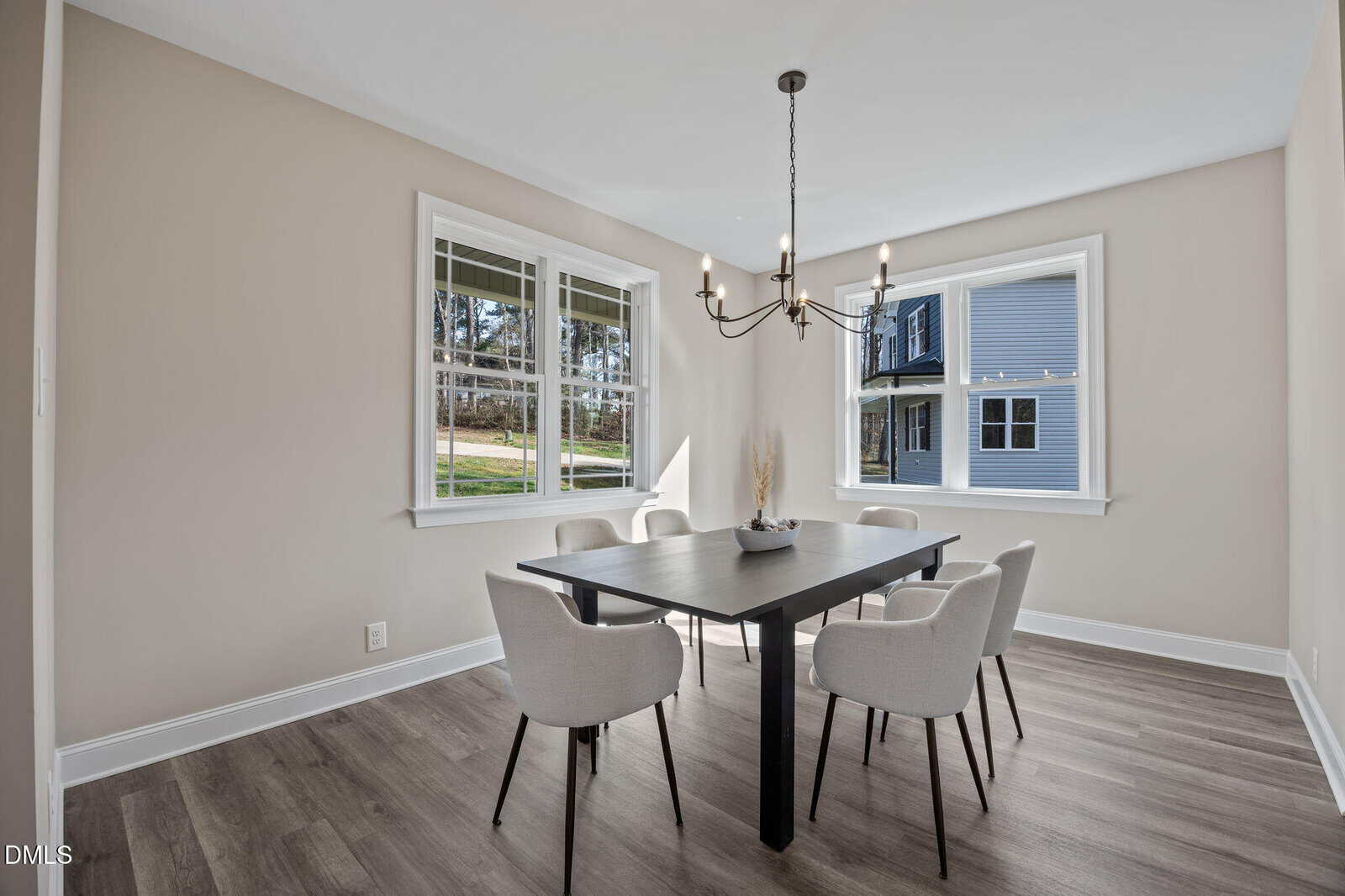 Modern dining room with light-colored walls, large windows, a black rectangular table, six white upholstered chairs, a decorative vase centerpiece, and a black chandelier. Natural light fills the space.