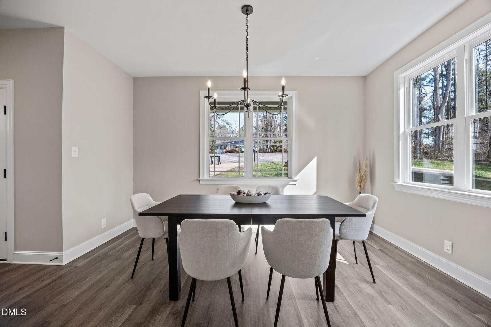 A dining room with a dark rectangular table, four white upholstered chairs, a bowl centerpiece, a modern chandelier overhead, large windows letting in natural light, and light-colored walls and flooring.