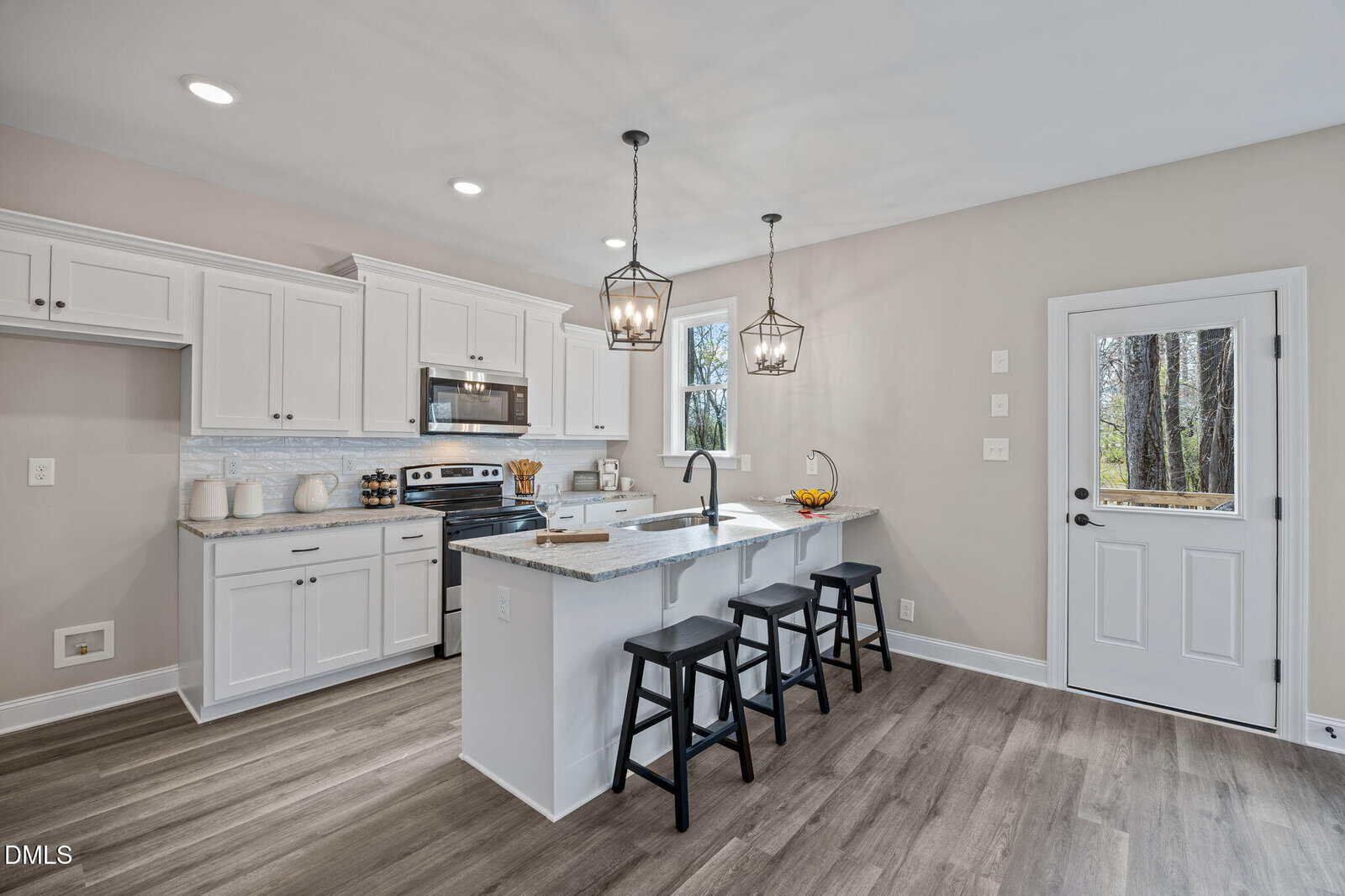 A modern kitchen with white cabinets, stainless steel appliances, a small island with three black stools, pendant lights, wood-style flooring, and a door leading outside with a window showing trees.