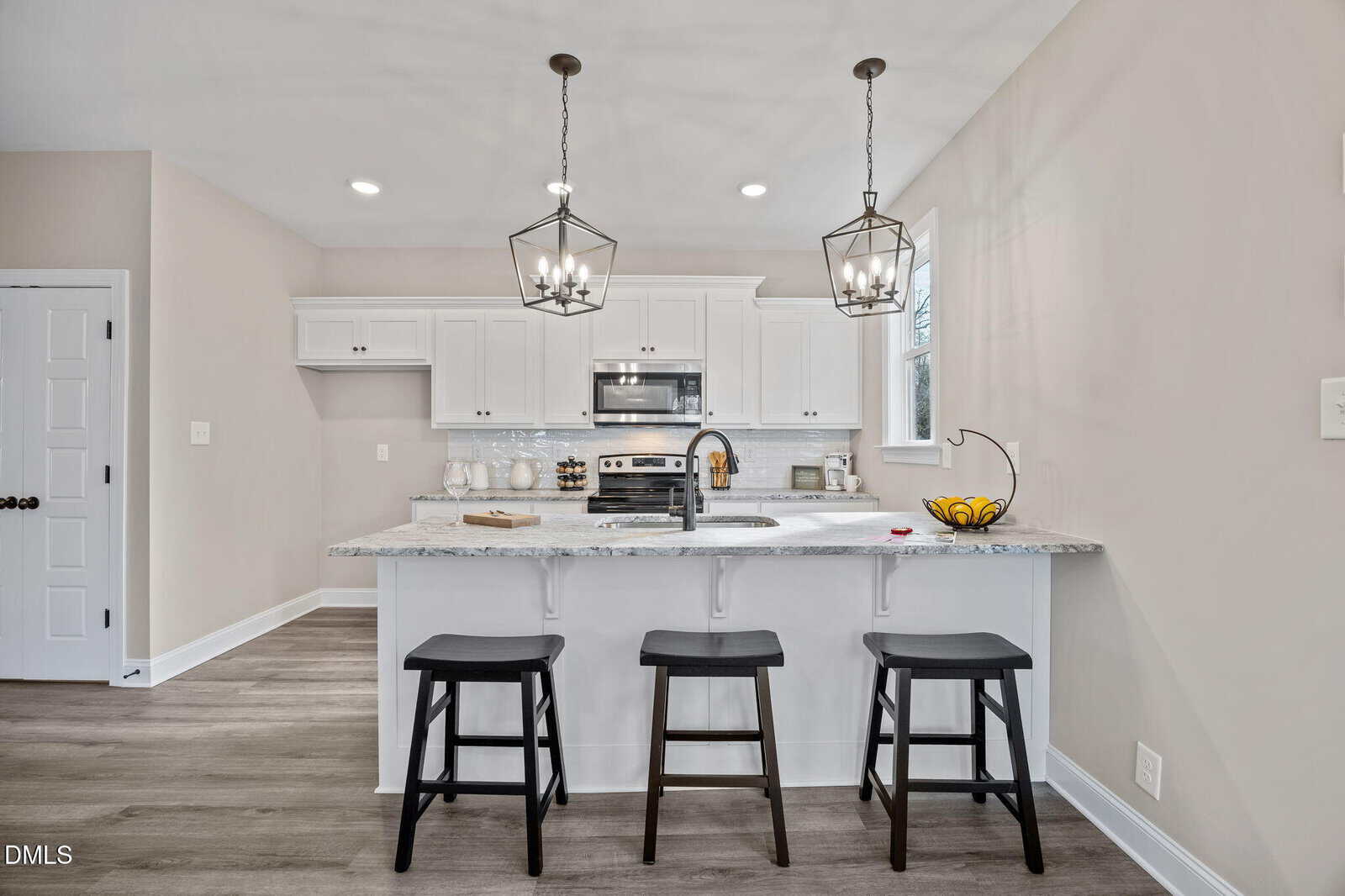 A modern kitchen with white cabinets, stainless steel appliances, and a marble countertop island with three black stools. Pendant lights hang above the island, and a bowl of fruit sits near the sink.