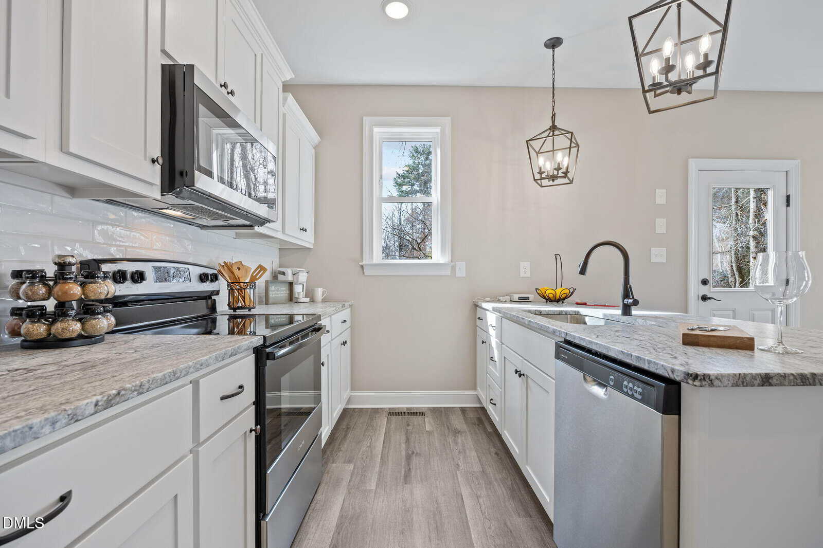A modern kitchen with white cabinets, stainless steel appliances, a gray stone countertop, a farmhouse sink, two geometric pendant lights, and a window showing trees outside. A bowl of bananas sits on the counter.