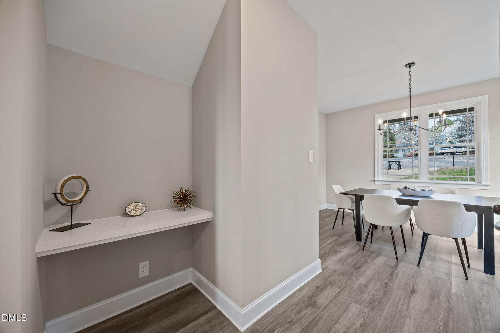 Modern dining area with a black table, four white chairs, a large window, and a black chandelier. Adjacent is a niche with a small white shelf displaying decorative objects against light-colored walls and wood flooring.