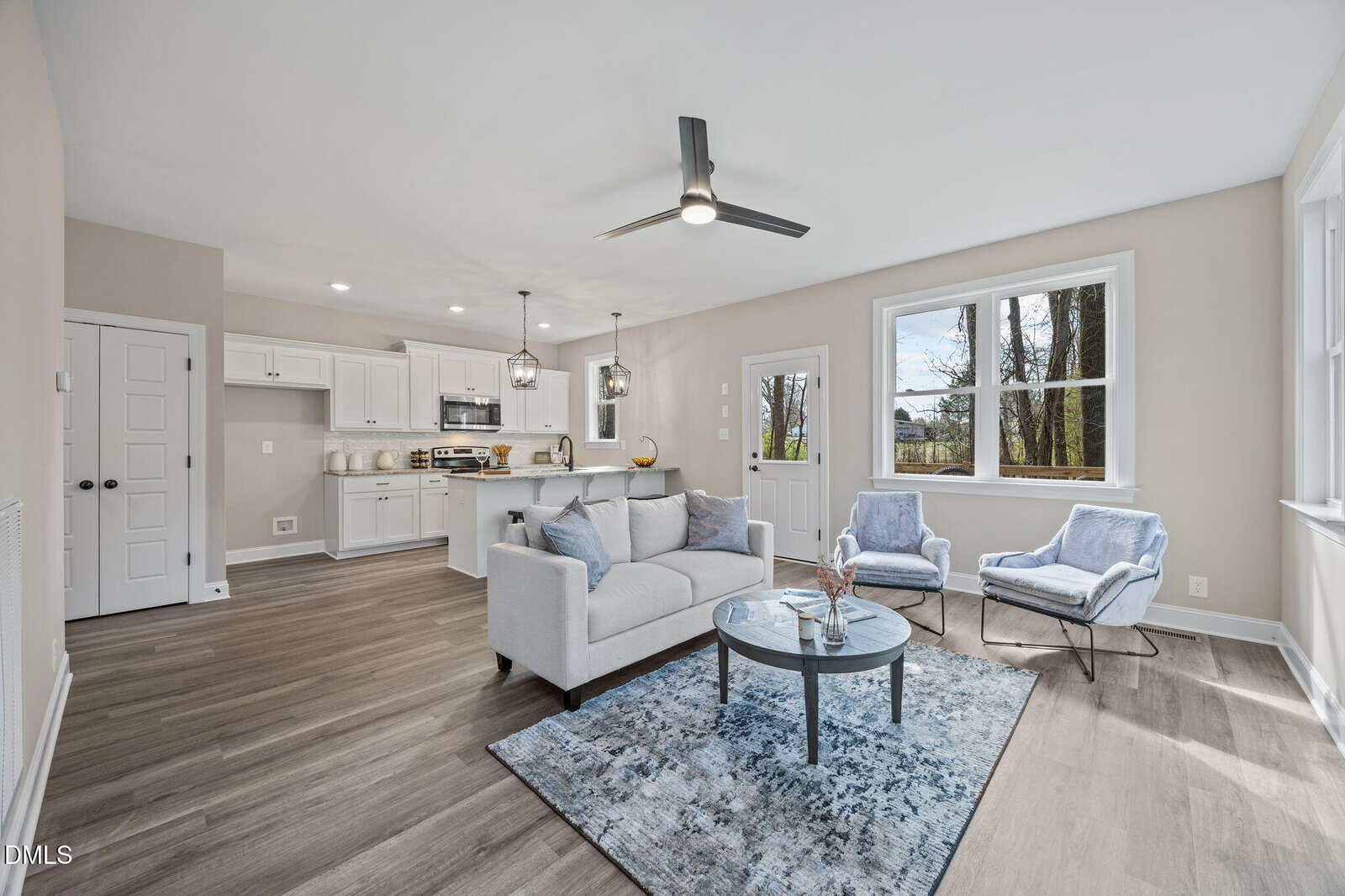 Modern open-concept living room and kitchen with light gray walls, large windows, a sofa, two armchairs, a round coffee table, and pendant lights over a kitchen island.
