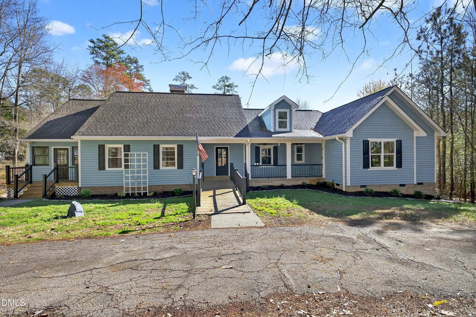 Charming single-story blue house with white trim at 810 Woodland Road in Creedmoor, featuring a welcoming front porch with a ramp, an American flag, gabled roof, black railings, lattice on the left side, a grassy yard, and surrounding trees under a clear sky.
