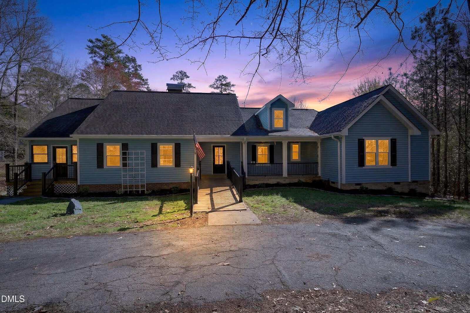 A single-story blue house with a dark roof is lit from within at dusk. A ramp and steps lead to the front door. There are trees in the background and a cracked driveway in the foreground.