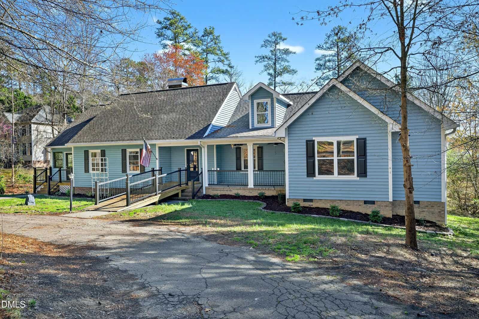 Single-story blue house with a gabled roof, front porch, ramp, and steps. American flag displayed by the entrance. Surrounded by trees and a partially shaded driveway. Sky is clear and sunny.