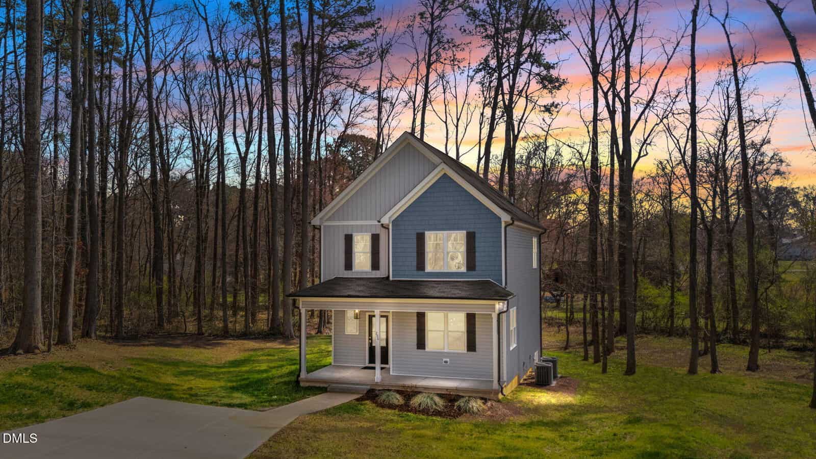 A two-story blue house with black shutters sits in a clearing surrounded by tall trees at sunset. The house has a covered front porch, dormer windows, and a driveway leading to the front.