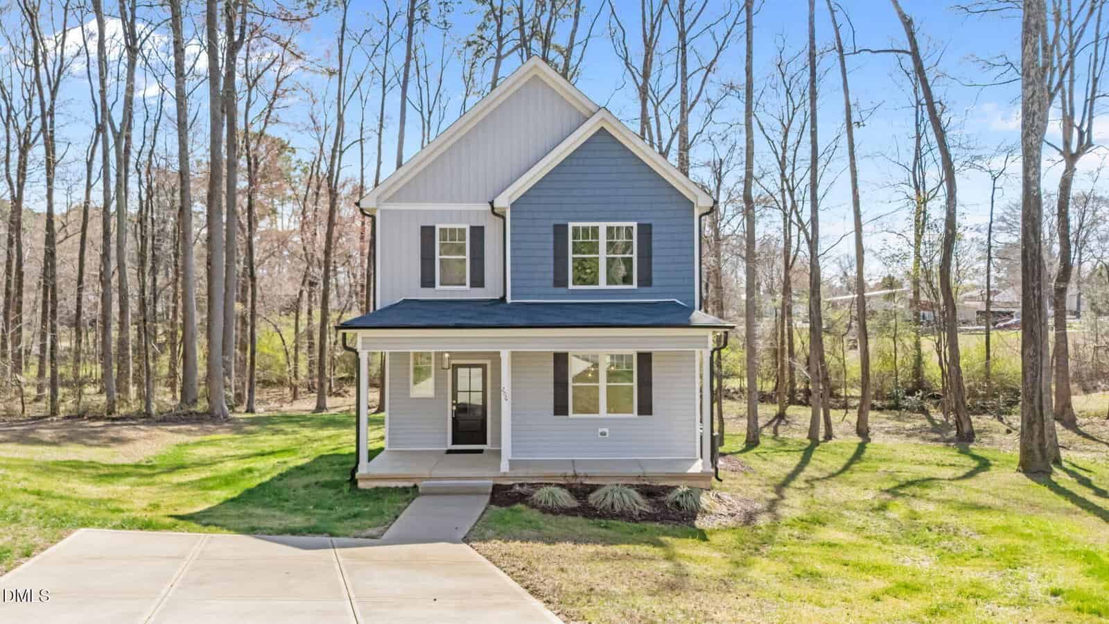 A two-story blue house with white trim sits in a grassy yard surrounded by tall, leafless trees. The house has a covered front porch, large windows, and a concrete driveway leading to a sidewalk.