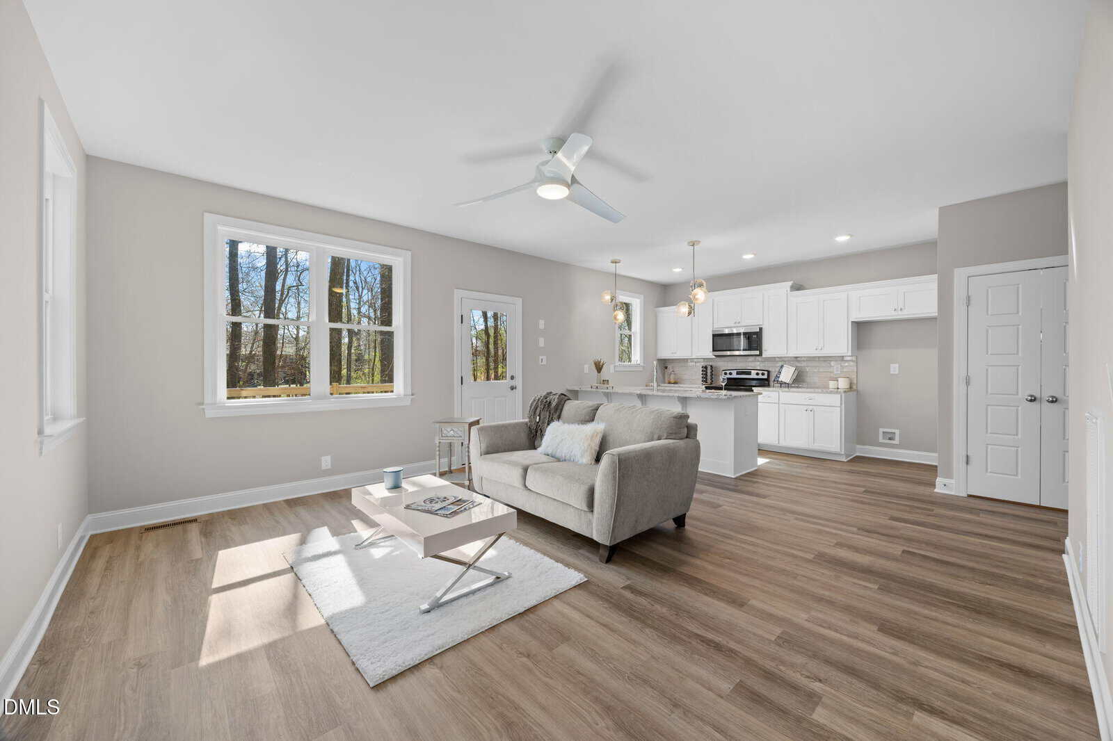 Bright, modern open-concept living room and kitchen with large windows, white walls, light wood flooring, gray sofa, coffee table, and overhead ceiling fan. Natural light fills the space.