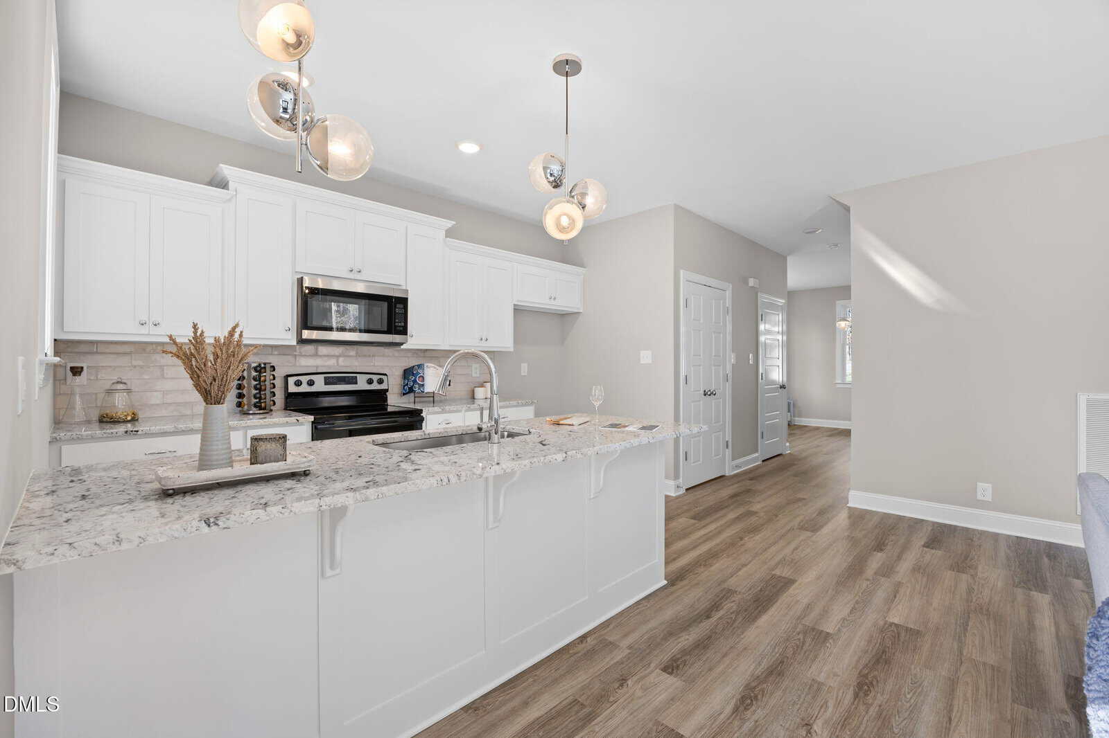 A modern kitchen with white cabinets, stainless steel appliances, and a marble countertop island with a sink. Pendant lights hang above the island, and wood-style flooring extends into an adjacent hallway.