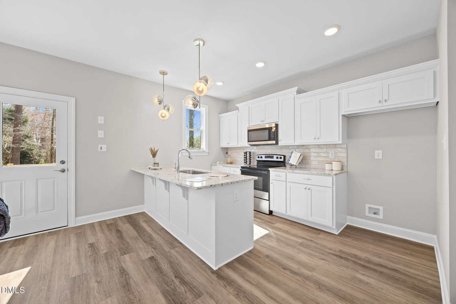 A modern kitchen features white cabinets, stainless steel appliances, a center island with a sink, pendant lighting, light countertops, and wood-look flooring. There is a window and a door letting in natural light.