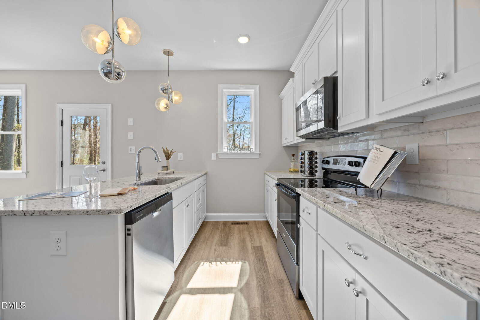 Modern kitchen with white cabinets, stainless steel appliances, marble countertops, pendant lights, a small window, light wood flooring, and a neutral backsplash. Natural light enters from the window and glass door.