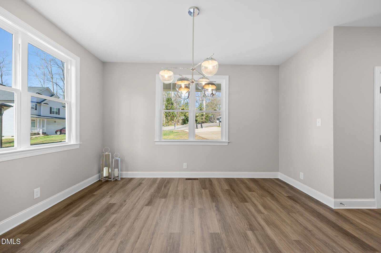 Dining area with wood floors, soft gray walls, modern chandelier, and large windows letting in natural light; two decorative lanterns sit in the corner.