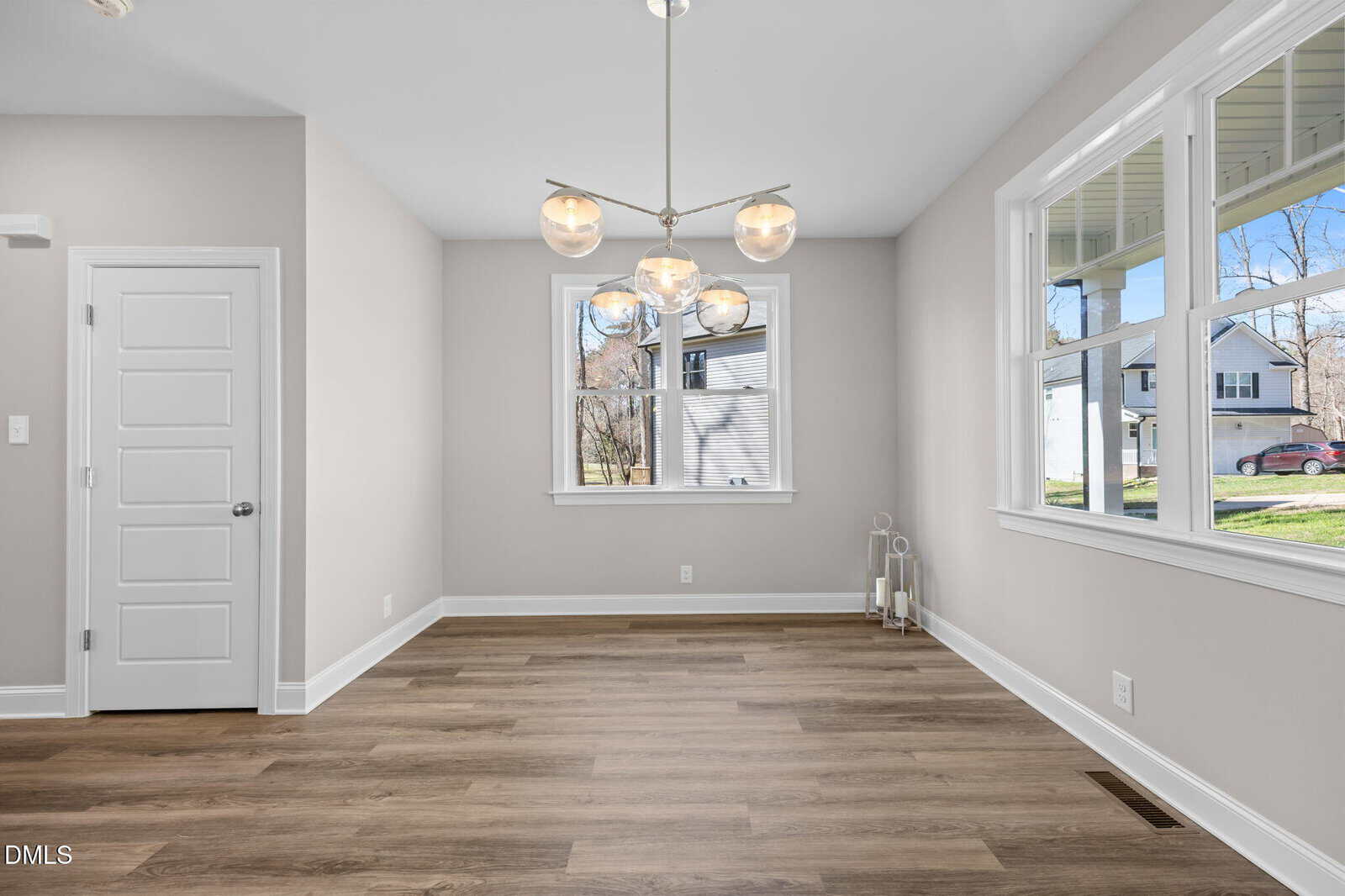 Bright, empty dining room with light wood flooring, modern chandelier, white walls, and large windows letting in natural light. A white door is on the left wall.