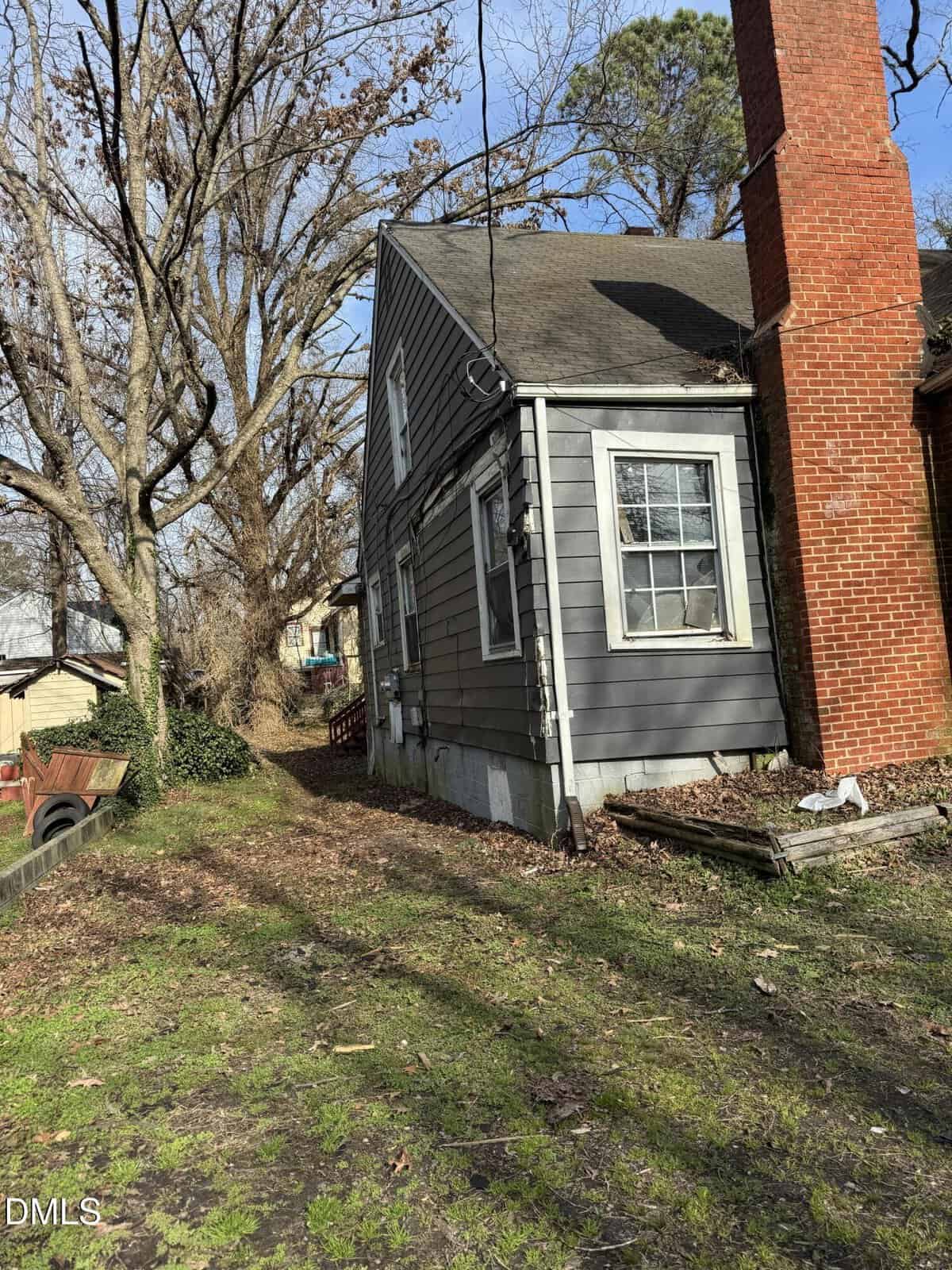 A gray, wood-sided house with a brick chimney sits next to a dirt driveway. Leafless trees and scattered leaves are visible, with some outdoor debris and a wagon near the side of the house.
