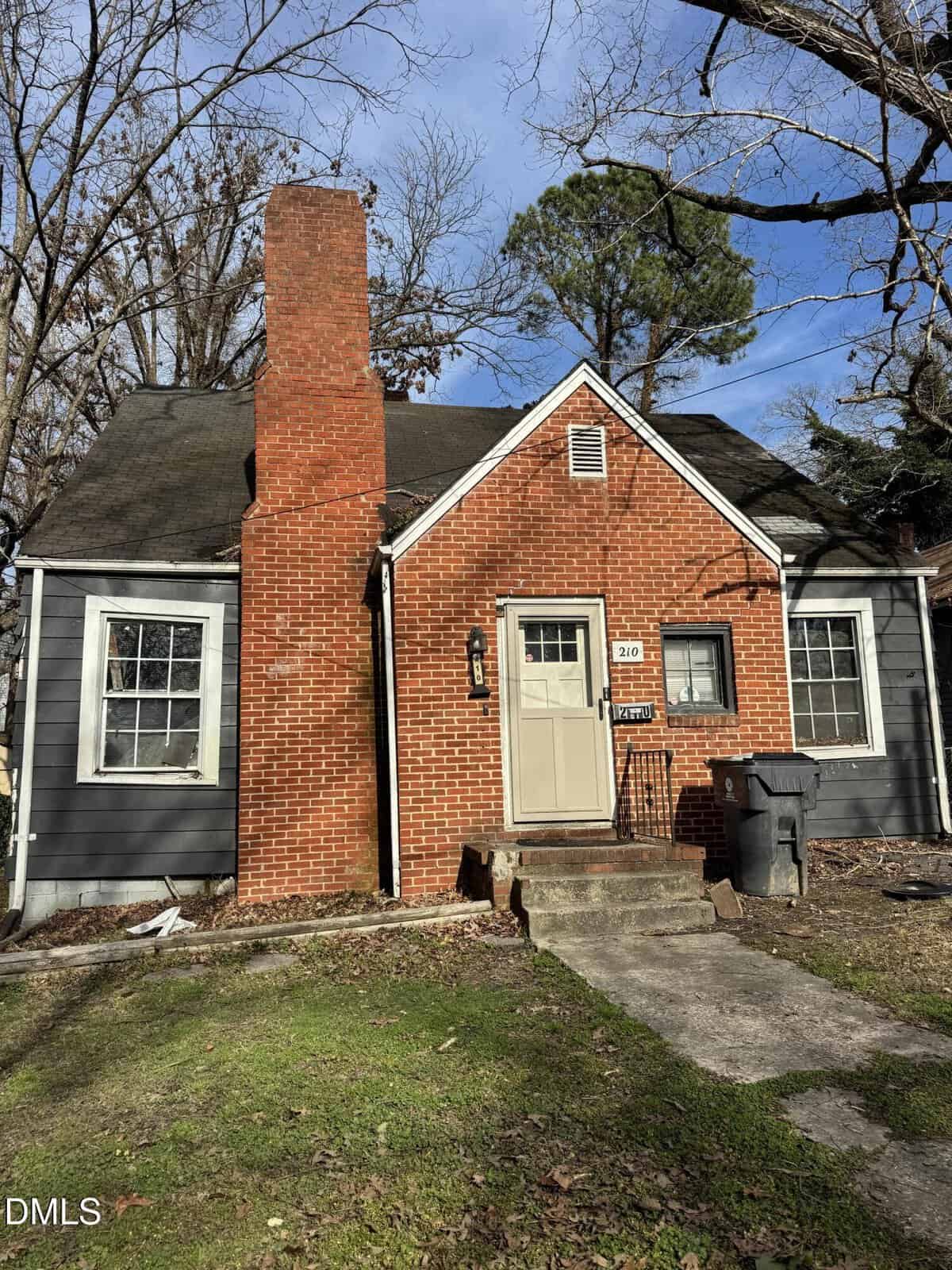 A small, older house with red brick chimney and entryway, gray siding, a shingled roof, and a brown front door. Two windows flank the entry. A trash bin sits at the right of the front steps. Trees are in the background.