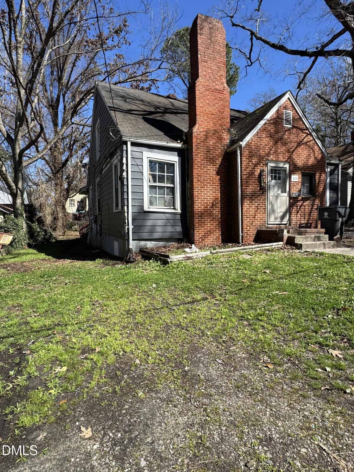 Small gray house with a red brick chimney and front steps, located at 210 Zollicoffer Avenue in Henderson. Surrounded by leafless trees, the yard features patchy grass and gravel, with a trash bin visible near the house. Bright sunlight casts clear shadows across the property.