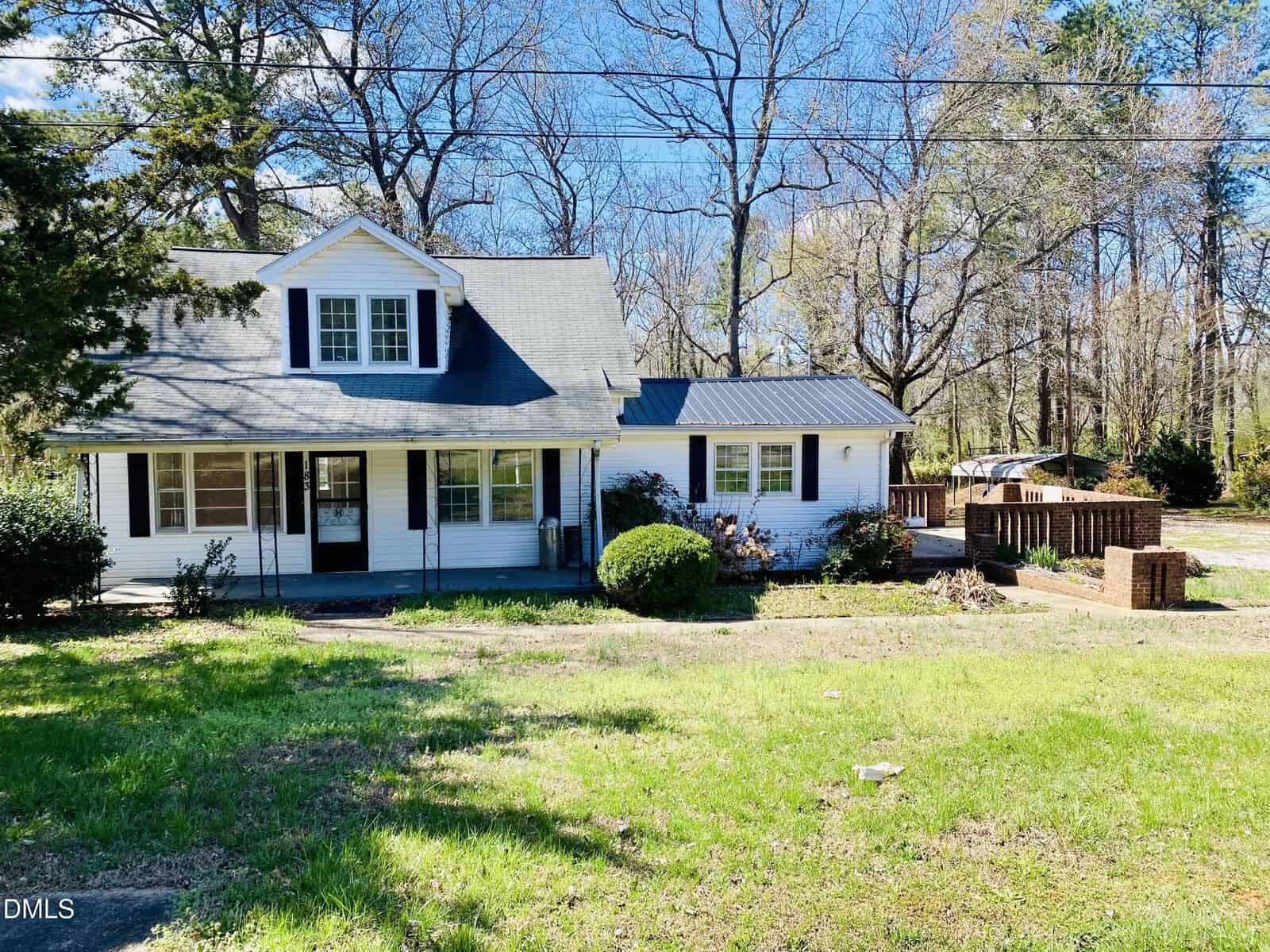 This charming white house with a black roof and dormer windows sits on 183 Red Hill Loop Road in Warrenton, surrounded by mature trees. There’s a brick patio area to the right and a grassy yard in front. The sky is clear and blue, with bare branches visible above the peaceful property.