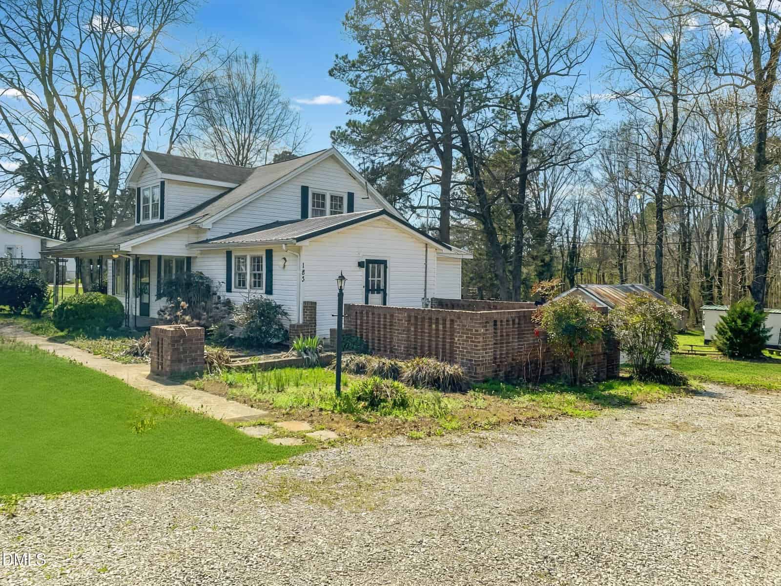 A white two-story house with a large front porch, surrounded by trees and shrubs, sits next to a brick garden enclosure and a gravel driveway under a clear blue sky.