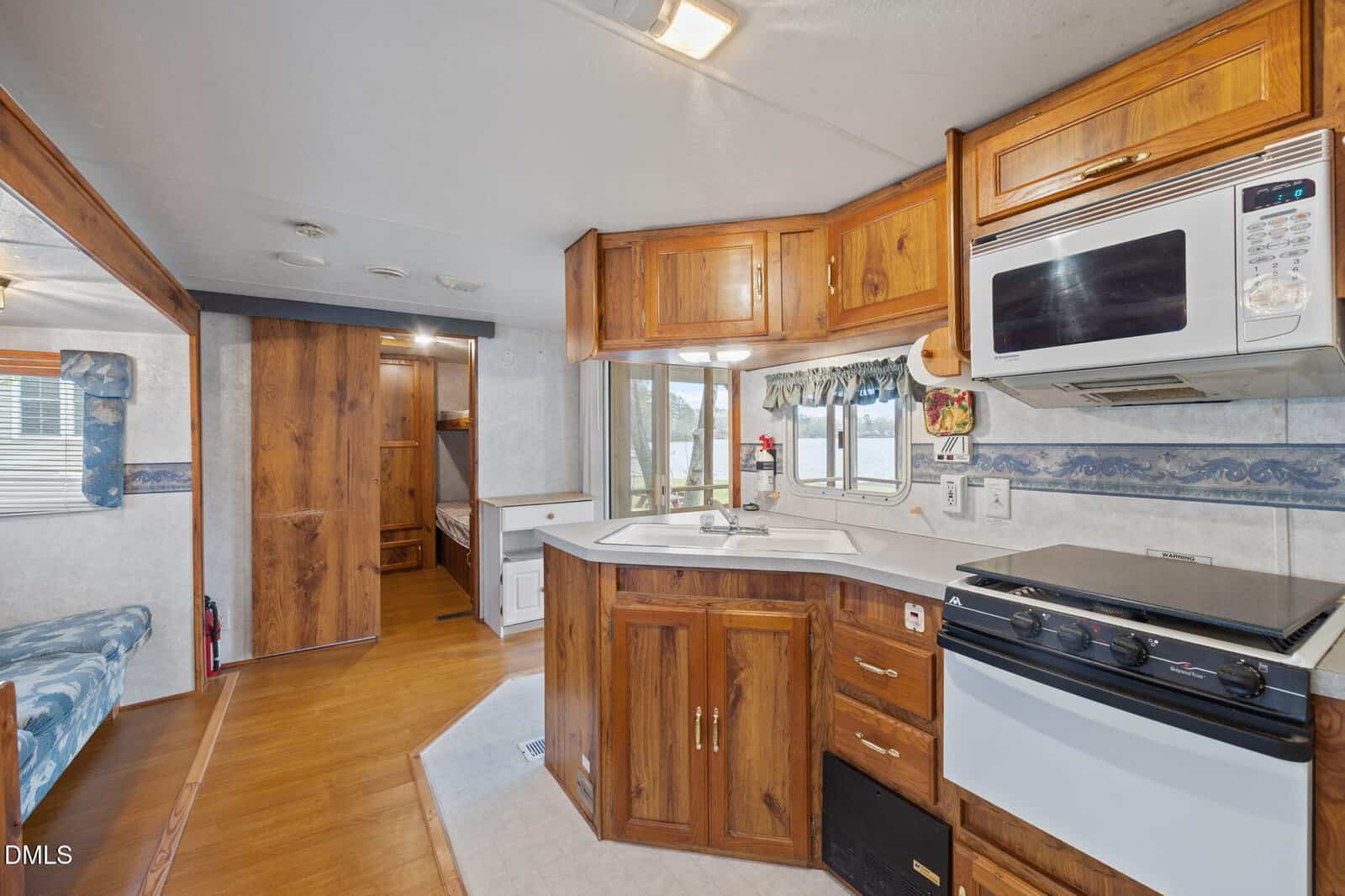 A compact RV kitchen features wooden cabinets, a microwave, a stove with an oven, a small sink, and a window with a valance. Adjacent spaces include a hallway, built-in bench, and visible sleeping area.