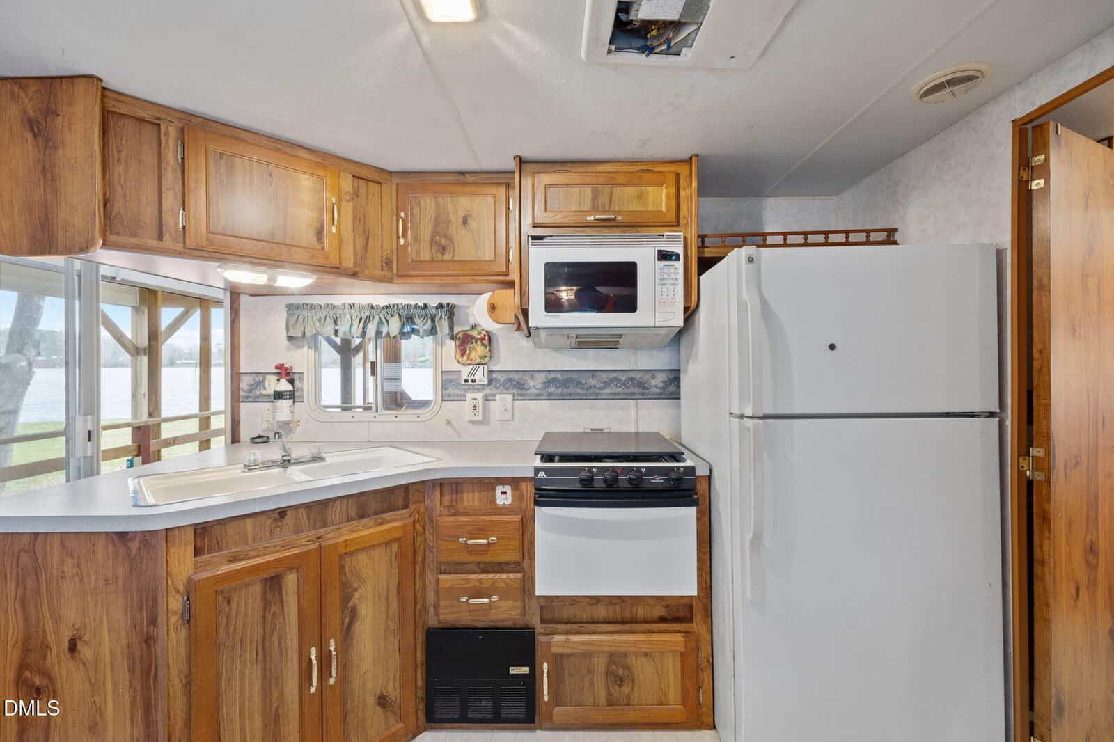 Small kitchen with wood cabinets, white refrigerator, stove, and microwave. Sink is under a window with a striped curtain. Blue patterned wallpaper strip runs along the back wall. Ceiling vent is partially open.