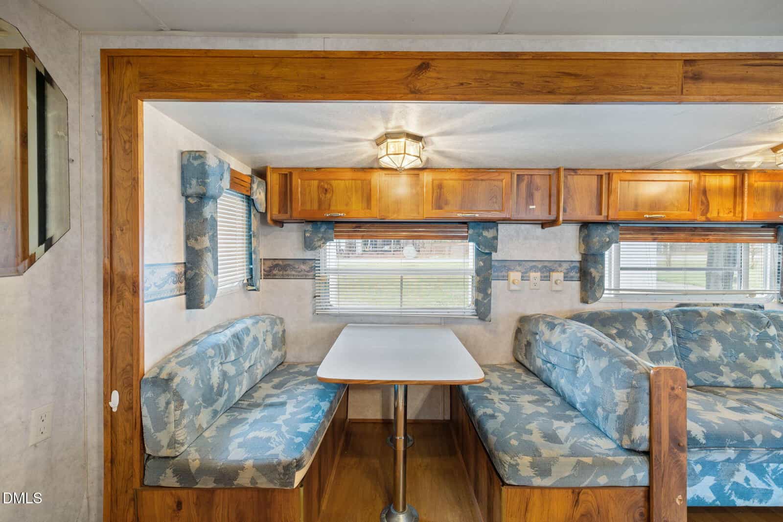 Interior view of an RV dining area with a table and two cushioned bench seats. There are wood cabinets above, blue patterned upholstery, windows with blinds, and a ceiling light fixture.